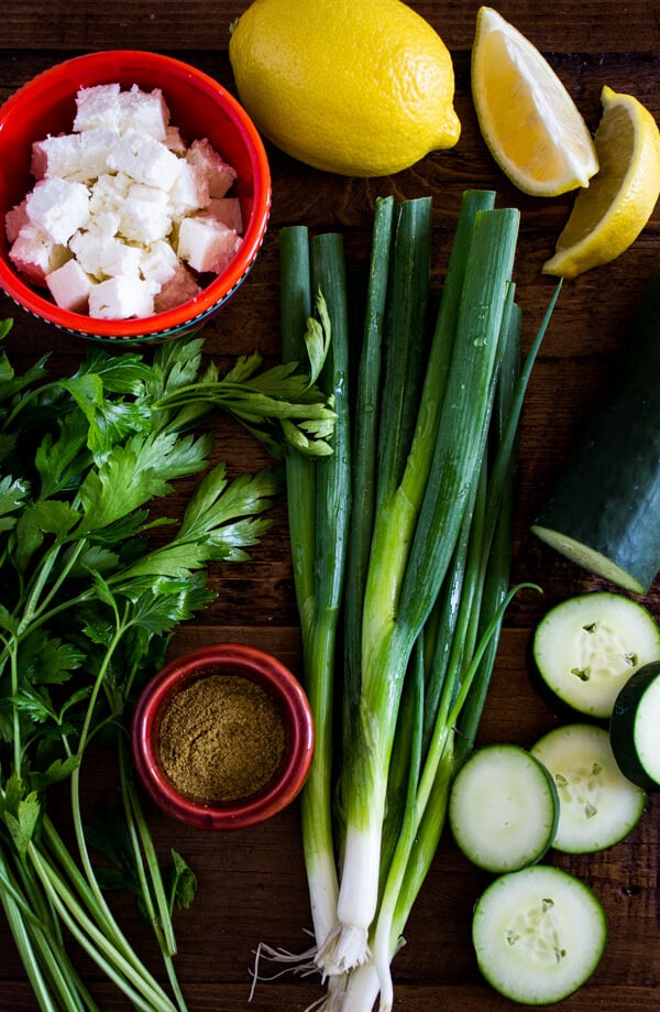 Lemons, cubes of feta cheese, Italian parsley, green onions, cucumber, and a small pinch bowl of cumin sit on a wood table.