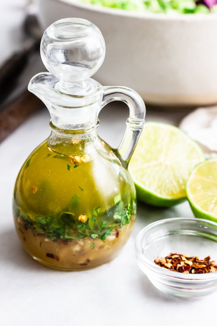 A small glass pouring jar with chili lime vinaigrette. A pinch bowl of red chili flakes and a few lime halves rest nearby.