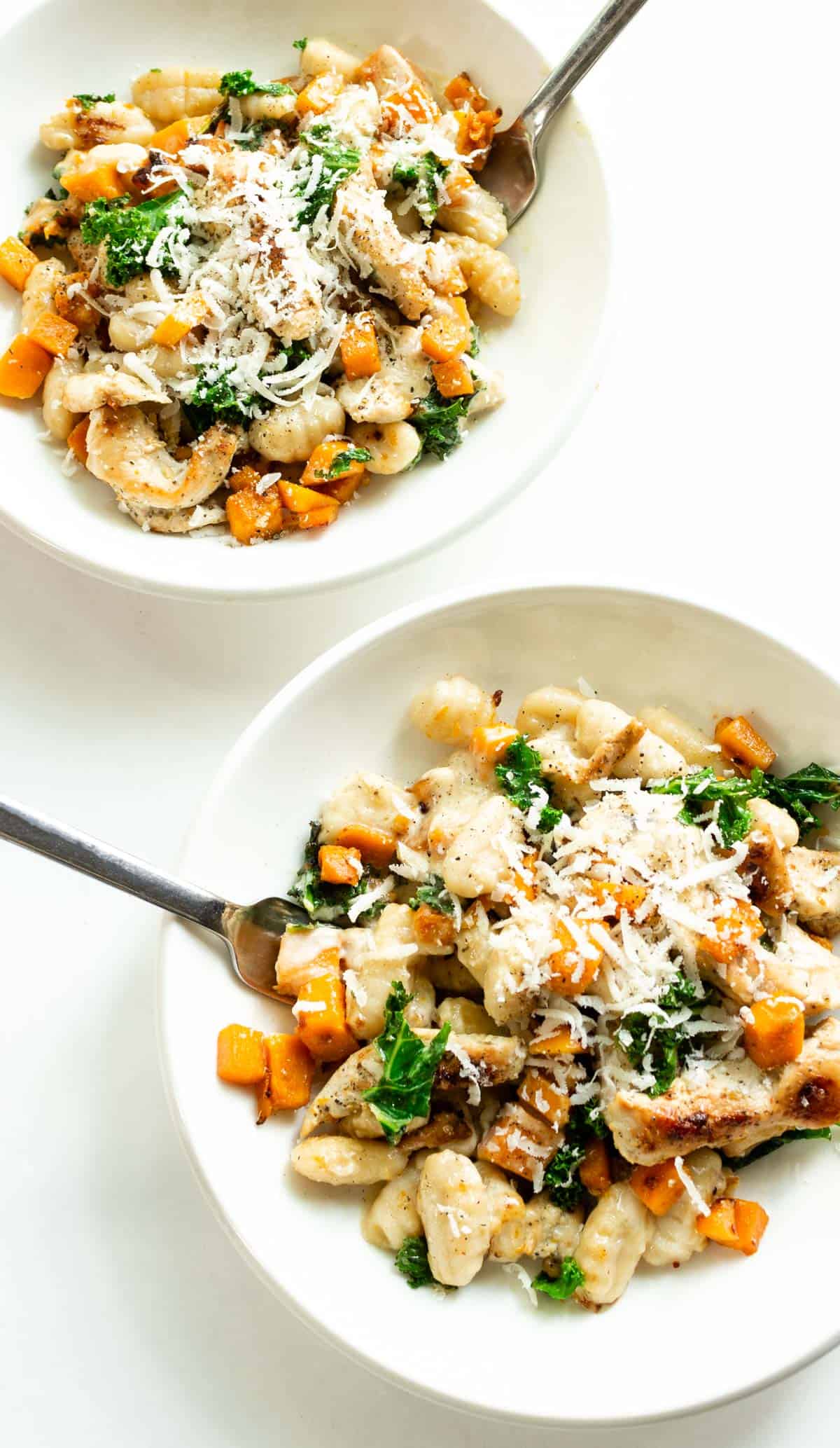 Overhead view of two white bowls on a white countertop with sliced grilled chicken with gnocchi, chopped butternut squash, and kale in a creamy sauce topped with grated parmesan.