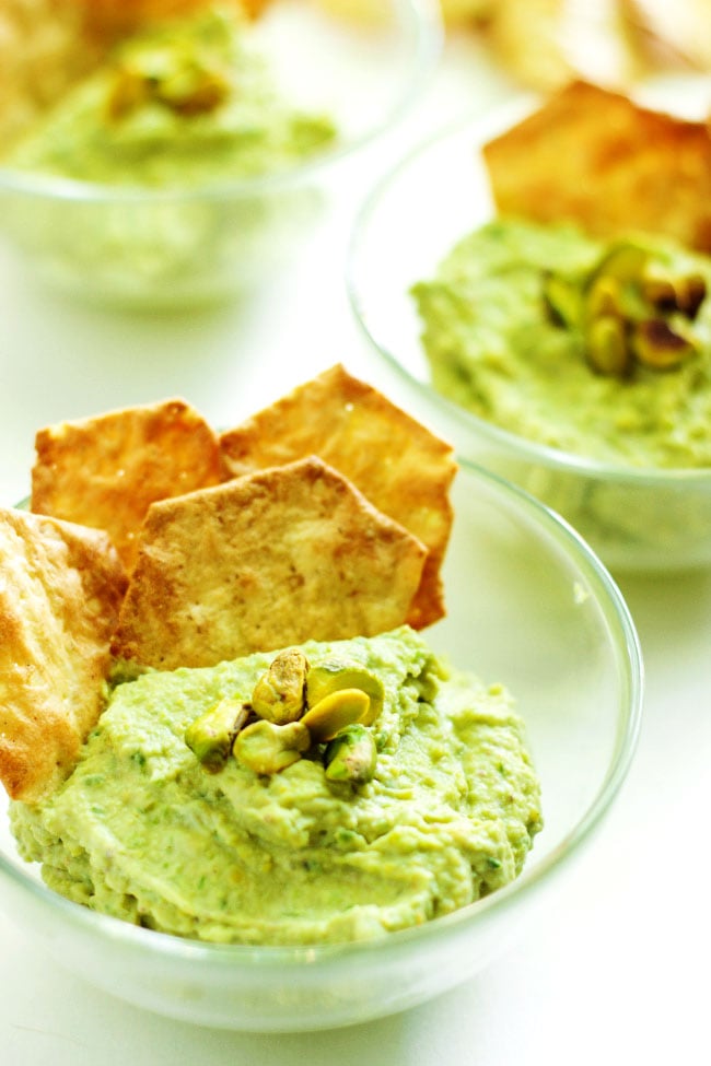 A close-up of several serving bowls of green garlic dips topped with pistachios and served with crackers.