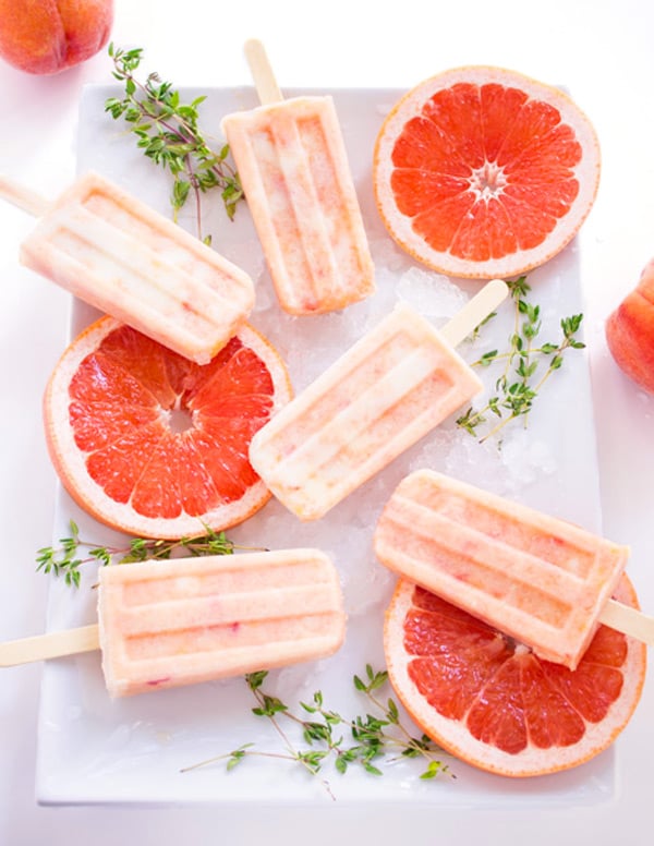 Grapefruit peach popsicles are laid out on a white plate with slices of fresh grapefruit and sprigs of fresh thyme.