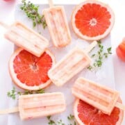 Grapefruit peach popsicles are laid out on a white plate with slices of fresh grapefruit and sprigs of fresh thyme.