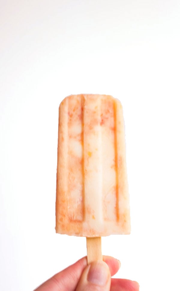 A hand is holding up a single grapefruit peach popsicle.