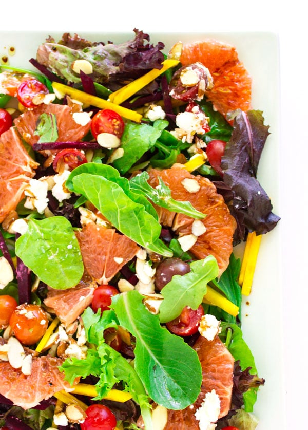 Slivered purple and yellow beets with green salad, cherry tomatoes, slivered almonds, feta cheese, and a drizzle of balsamic vinaigrette on a white, square platter.