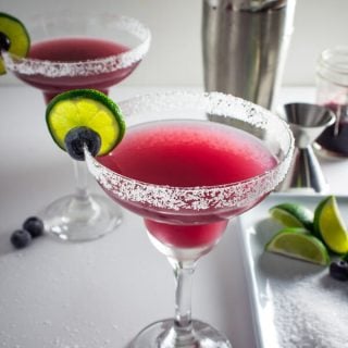 A Blue Q Margarita in a cocktail glass with a salted rim. and garnish wish a lime wheel and a blueberry. Silver cocktail shaker, limes wedges, salt, blueberries, and blue q sauce in the background.