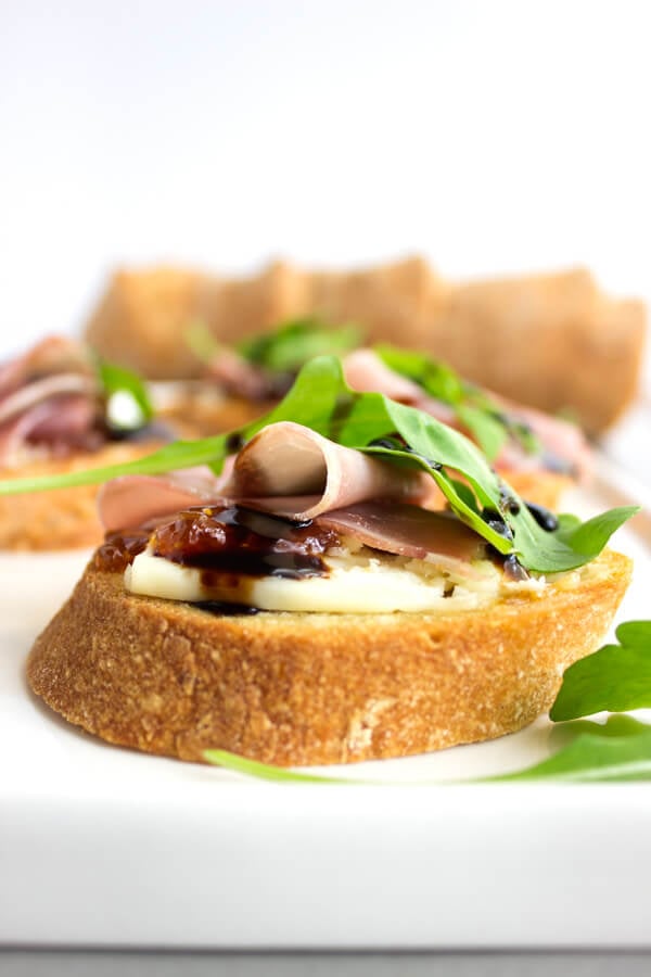 A slice of gluten-free crostini with a schmear of fig jam and port salut cheese topped with a salty slice of prosciutto, peppery arugula, and drizzle of balsamic.
