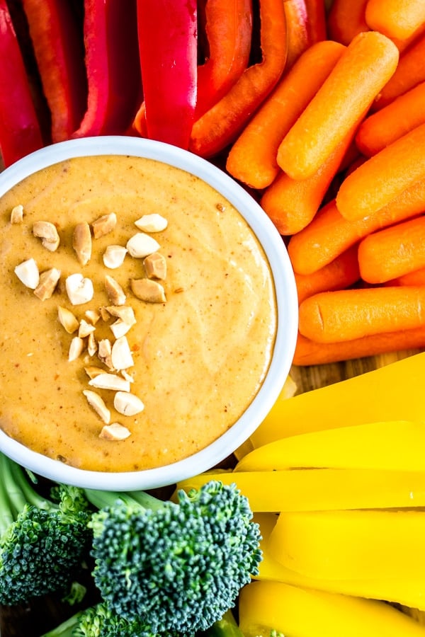 A dish of peanut sauce surrounded by red bell peppers, organ carrots, yellow bell peppers, and green broccoli.