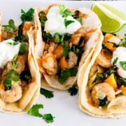 Shrimp tacos with onions, bell peppers, and sautéed kale in a corn tortilla with lime wedges on the side.