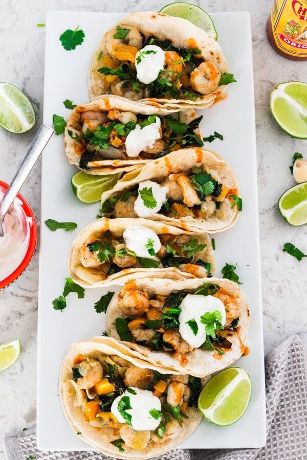 A long rectangular white plate with six shrimp tacos served with dollops of sour cream on top and drizzled with hot sauce. Fresh lime wedges are scattered around the plate along with a bottle of hot sauce and a red bowl of sour cream.
