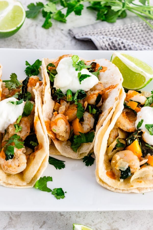 Three shrimp tacos with onions, bell peppers, and sautéed kale. Finished with a big squeeze of lime, a drizzle of hot sauce, fresh cilantro, and a dollop of sour cream all wrapped up in a warm soft corn tortilla with extra lime wedges on the side.