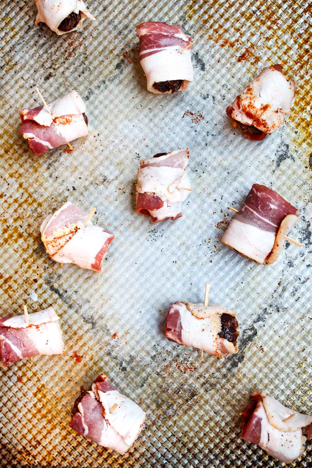 Dates have been wrapped in bacon with chili powder and are spread on a baking sheet ready to be baked.