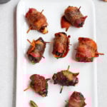 Chili bacon wrapped dates are served on a white rectangular plate. Chili powder is in a small dish off to the side.