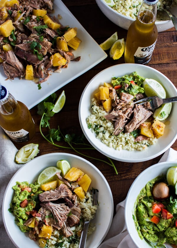 A tabletop with a large serving dish of pineapple pulled pork. A big bowl of guacamole. A bowl of cilantro lime cauliflower rice. And two plates loaded up with food ready to be eaten. Lime wedges and a couple of Corona's nearby.