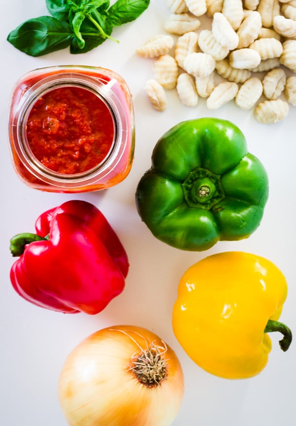 Tri-colored bell peppers, an onion, a jar of marinara, gnocchi, and fresh basil are set out on a white counter.