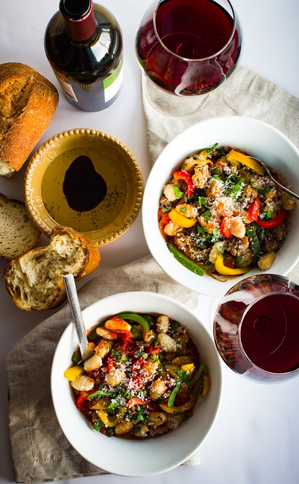 Two bowls of gnocchi marinara with sausage and peppers are paired with a bottle of red wine. Slices of crusty bread with oil & balsamic for dipping are nearby.