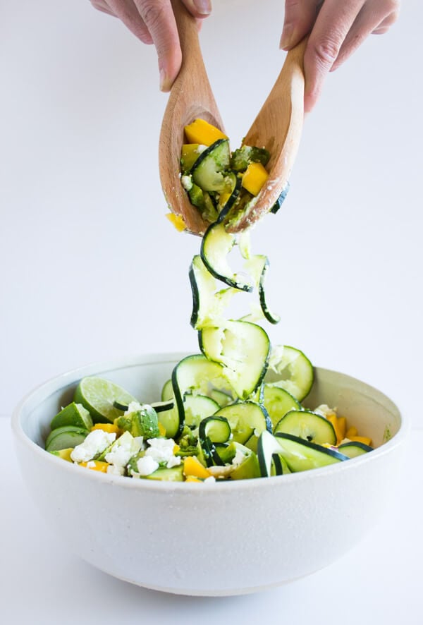 Two hands are reaching into a spiralized cucumber summer salad with serving spoons.