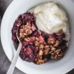 A close-up view of a bowl of blackberry apple hazelnut crisp with a scoop of vanilla ice cream nestled in.