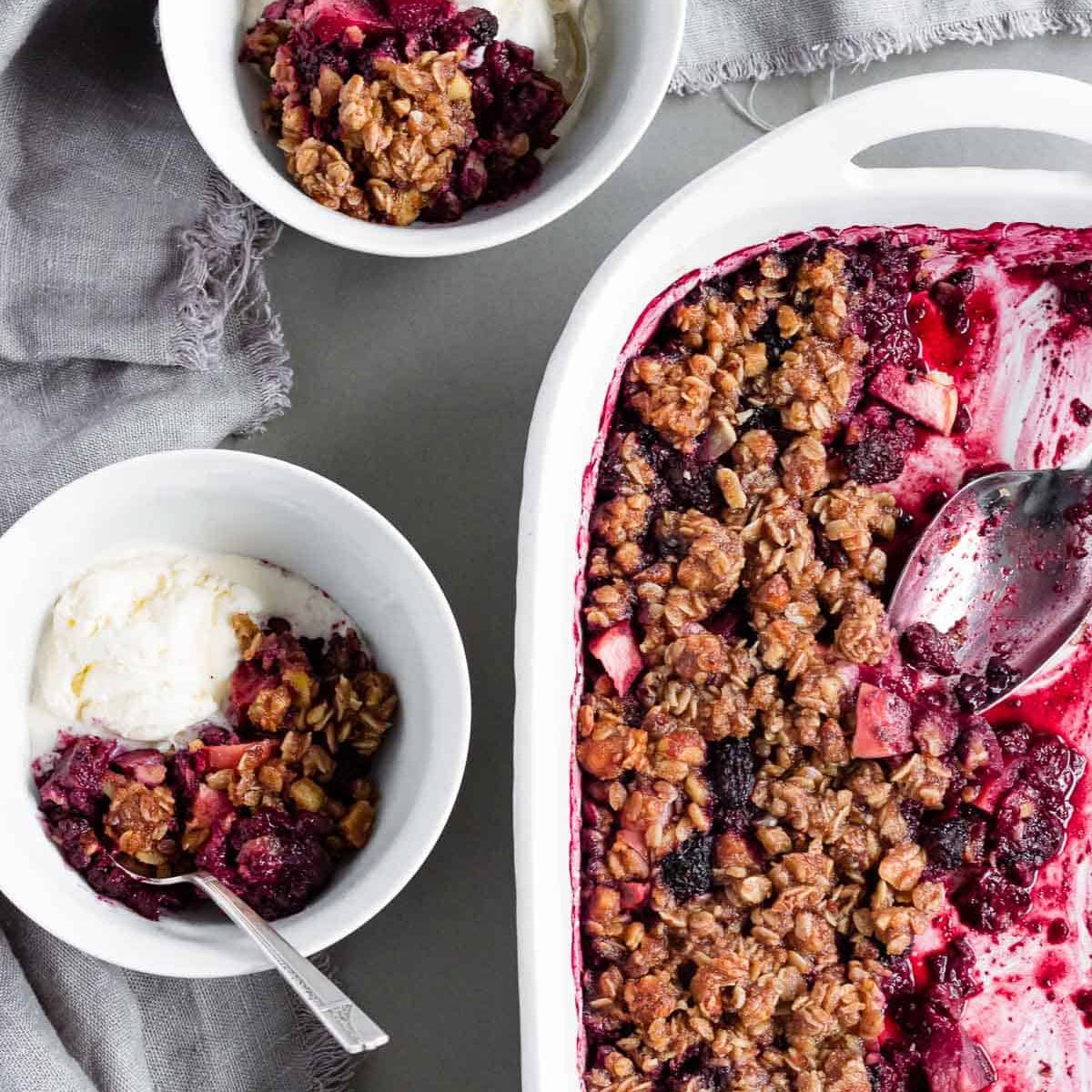 A large baking dish of healthier blackberry apple hazelnut crisp alongside two bowls of the crisp served with vanilla ice cream.