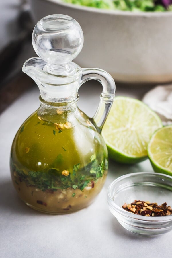 A small glass pouring jar with chili lime vinaigrette. A pinch bowl of red chili flakes and a few lime halves rest nearby.
