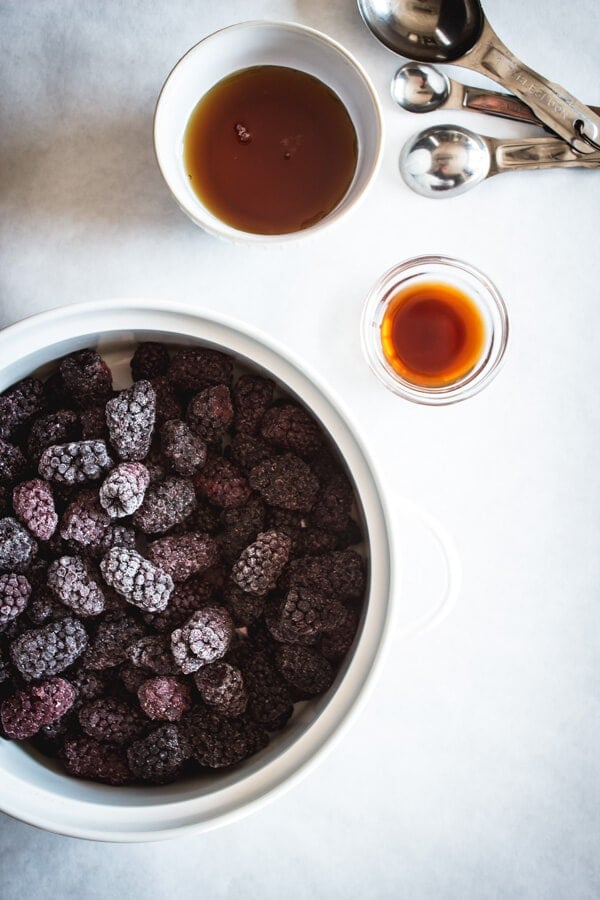 A baking dish with frozen Oregon Marionberries, two small bowl of maple syrup and vanilla, and silver measuring spoons nearby.