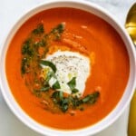 A white bowl of roasted red pepper tomato soup topped with burrata, basil, and a swirl of pesto.