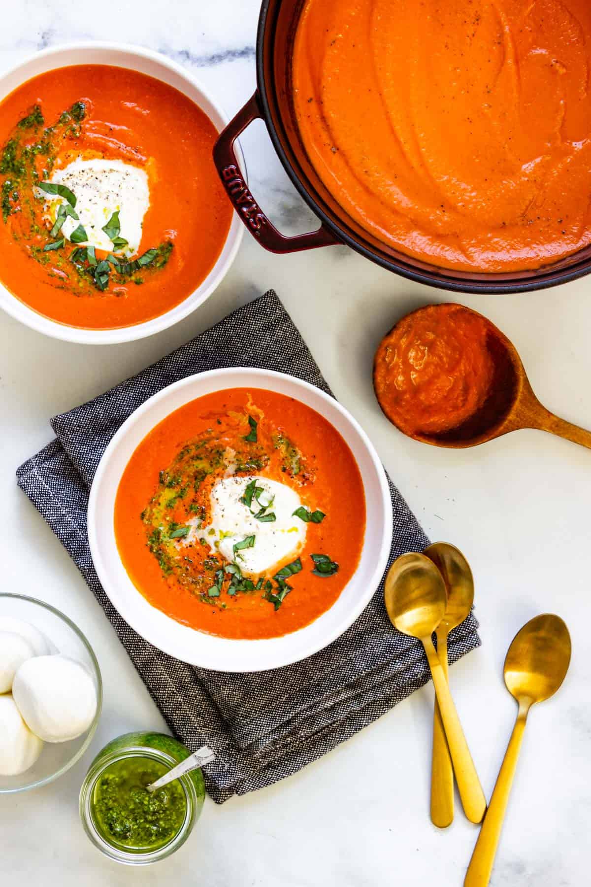 A pot and two bowls of soup, a bowl of burrata, and several gold spoons sit on a counter.