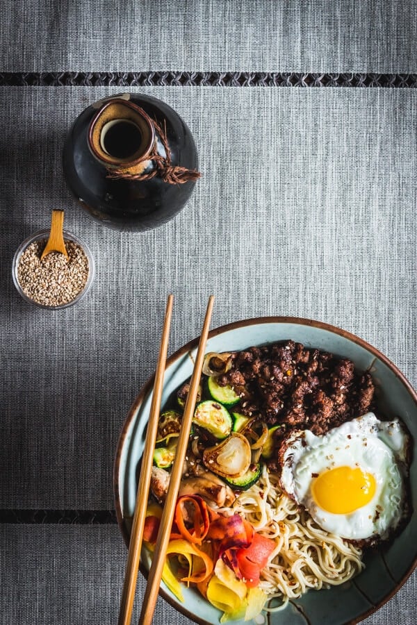 A bowl of ramen with veggie, beef, and fried egg sit on a grey tablecloth.