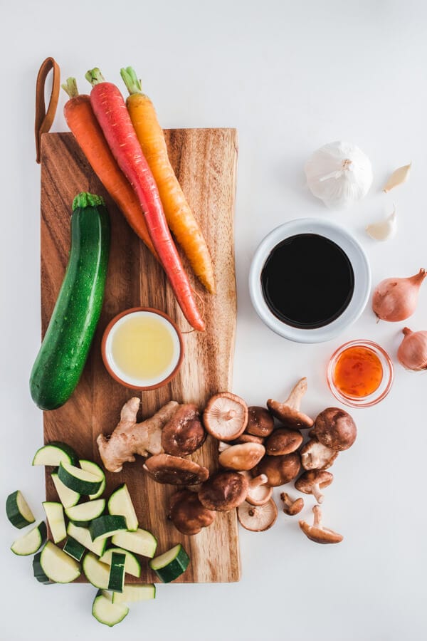 Zucchini, rainbow carrots, shiitake mushrooms, fresh ginger root sit on a cutting board. Sauce ingredients sit alongside.