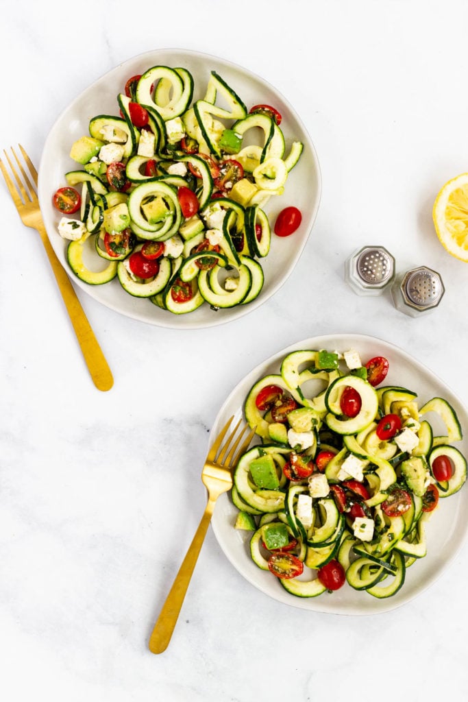 Two plates with a serving of zucchini, tomato, basil, feta salad. Two gold forks sit alongside.
