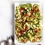 A beautiful spiralized zucchini salad with grape tomatoes, feta, and avocado tossed together on a square white serving dish.