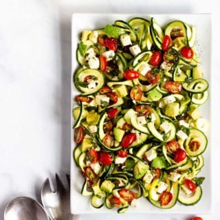 A beautiful spiralized zucchini salad with grape tomatoes, feta, and avocado tossed together on a square white serving dish.