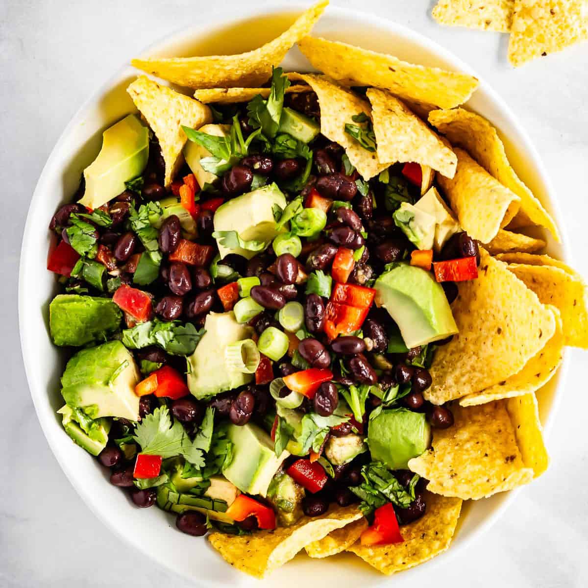 A bowl of avocado black bean salsa with tortilla chips tucked into the sides and scattered on the countertop.