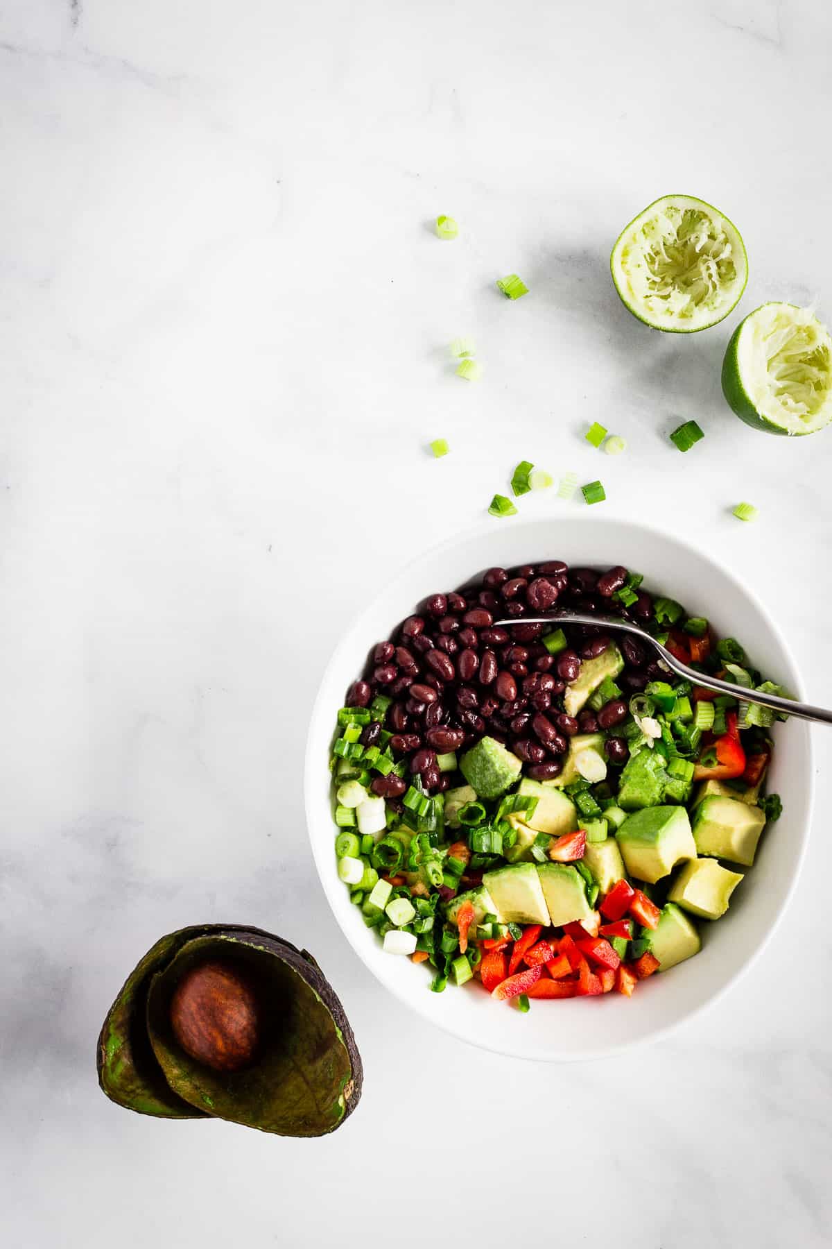 Combining ingredients for avocado black bean salsa in a white bowl. Avocado peels and squeezed limes sit nearby.