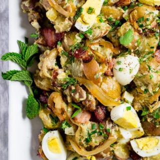 A close-up of view of potato salad with caramelized onions, slices of hard boiled eggs, chunks of salty crispy bacon, and fresh herbs.