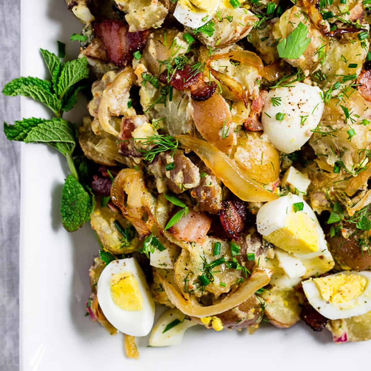 Potato salad with caramelized onions and fresh herbs is served obn a white rectangular serving dish. Garnished with fresh mint, dill, and chives.