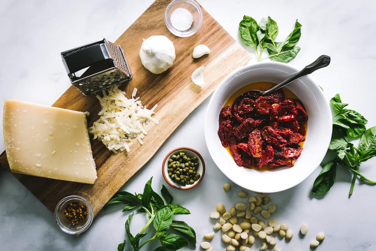 Ingredients to make homemade sun-dried tomato pesto are laid out on a marble counter and wood board.