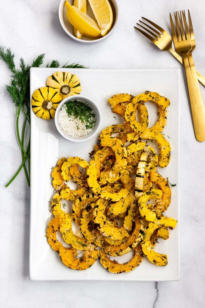 A white platter with roasted delicata squash with Parmesan and dill. A small dish of lemon wedges are served nearby.