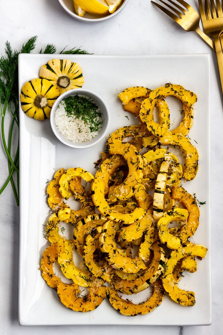 Roasted delicata squash served on a white platter with finely grated Parmesan, fresh dill, and lemon wedges.