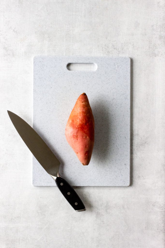 A sweet potato is resting on a cutting board with a knife.