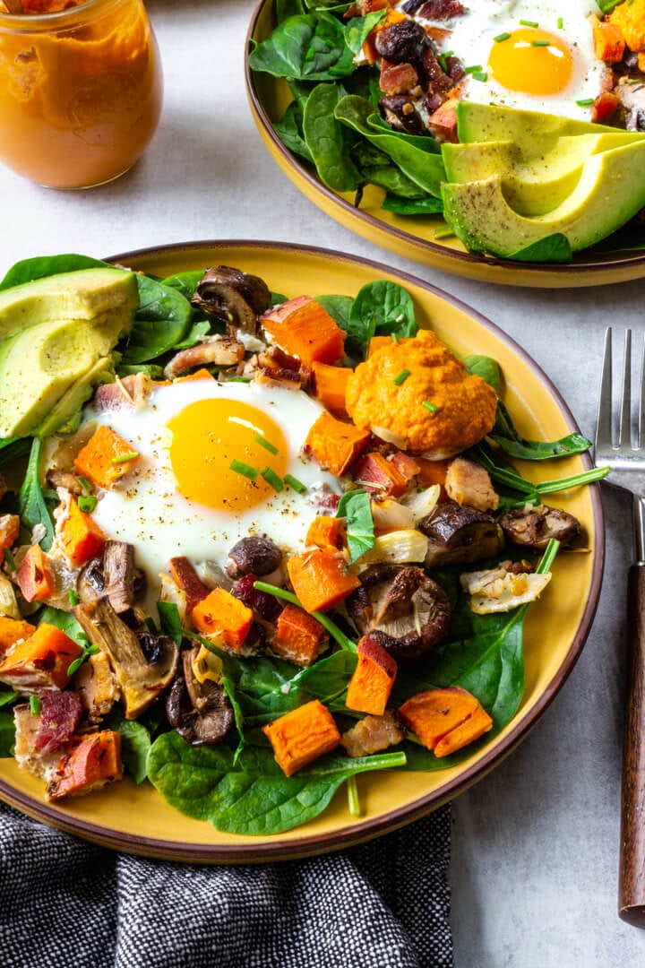 Sheet pan sweet potato hash with baked eggs, sliced avocado, and homemade romesco are served over baby spinach.