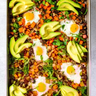 A sheet pan of roasted vegetables topped with baked eggs, avocado slices, and served over spinach.