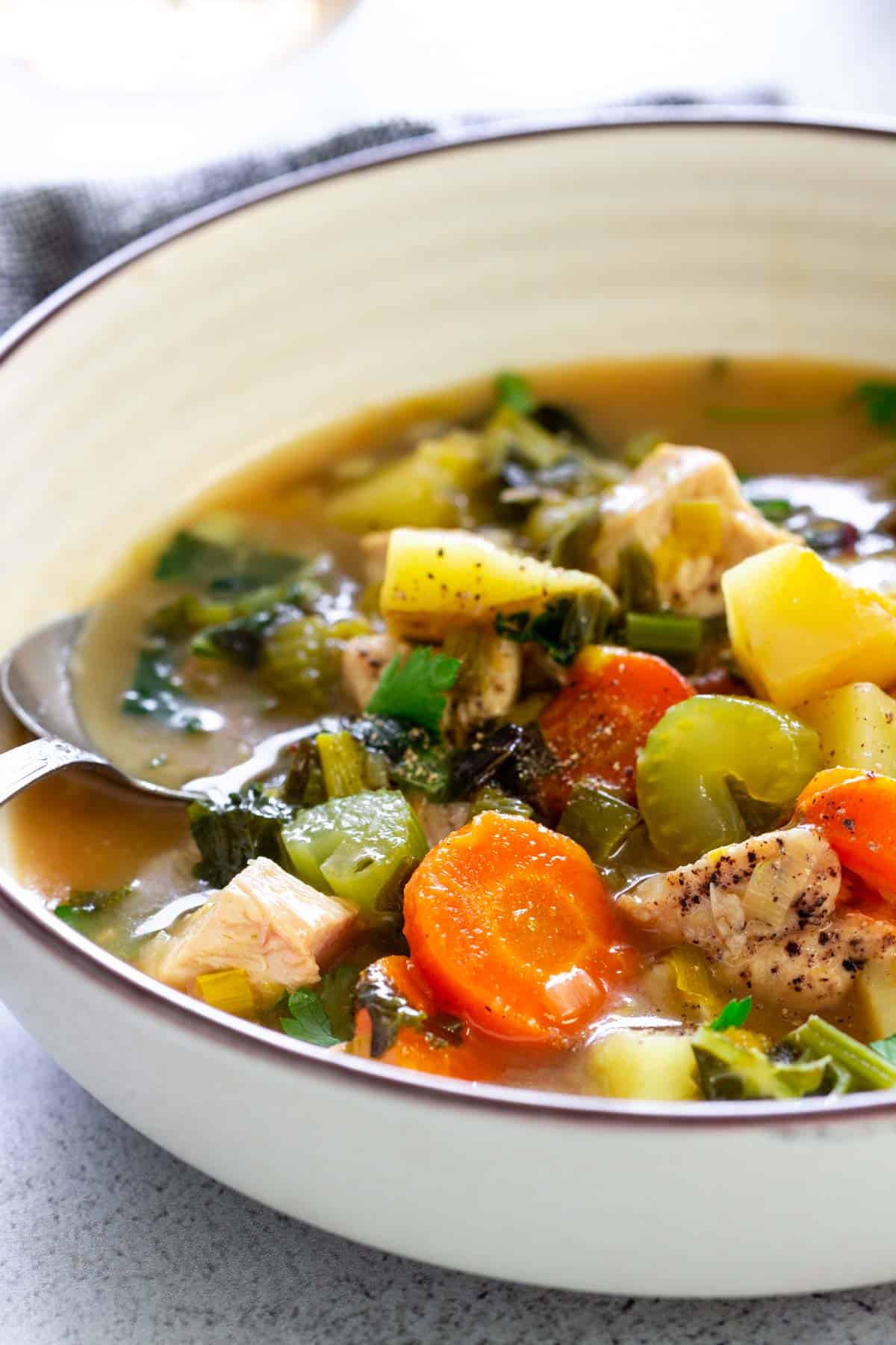 A close-up view of chicken soup loaded with chunks of chicken, carrots, celery, potatoes, fresh herbs, and winter greens.