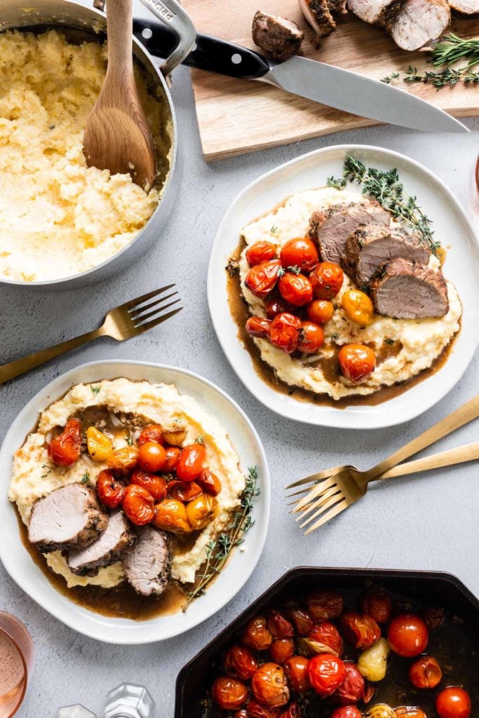 A table set with a pot of creamy polenta, a cutting board with sliced herb-crusted pork tenderloin, a skillet of roasted cherry tomatoes, and two plates with a serving of each.
