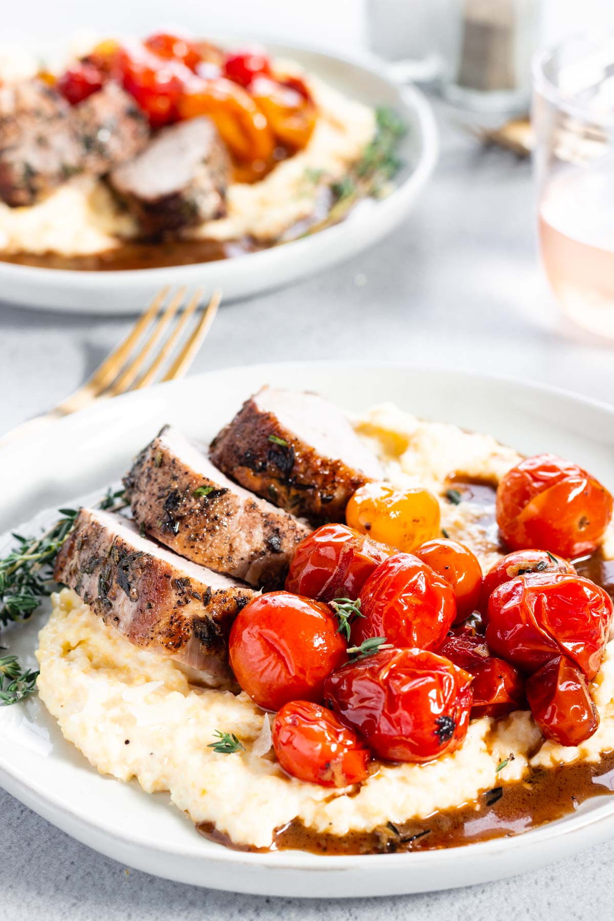 A plate with several slices of herb-crusted pork tenderloin served over creamy polenta and roasted cherry tomatoes.