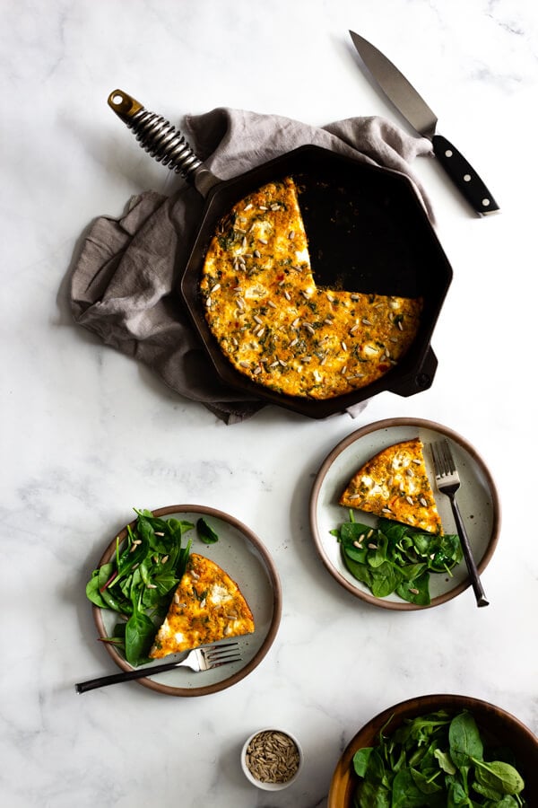 A sausage, kale, and sun-dried tomato pesto frittata baked in a cast iron skillet. Two ceramic plates with a serving of frittata and a side salad sit nearby.
