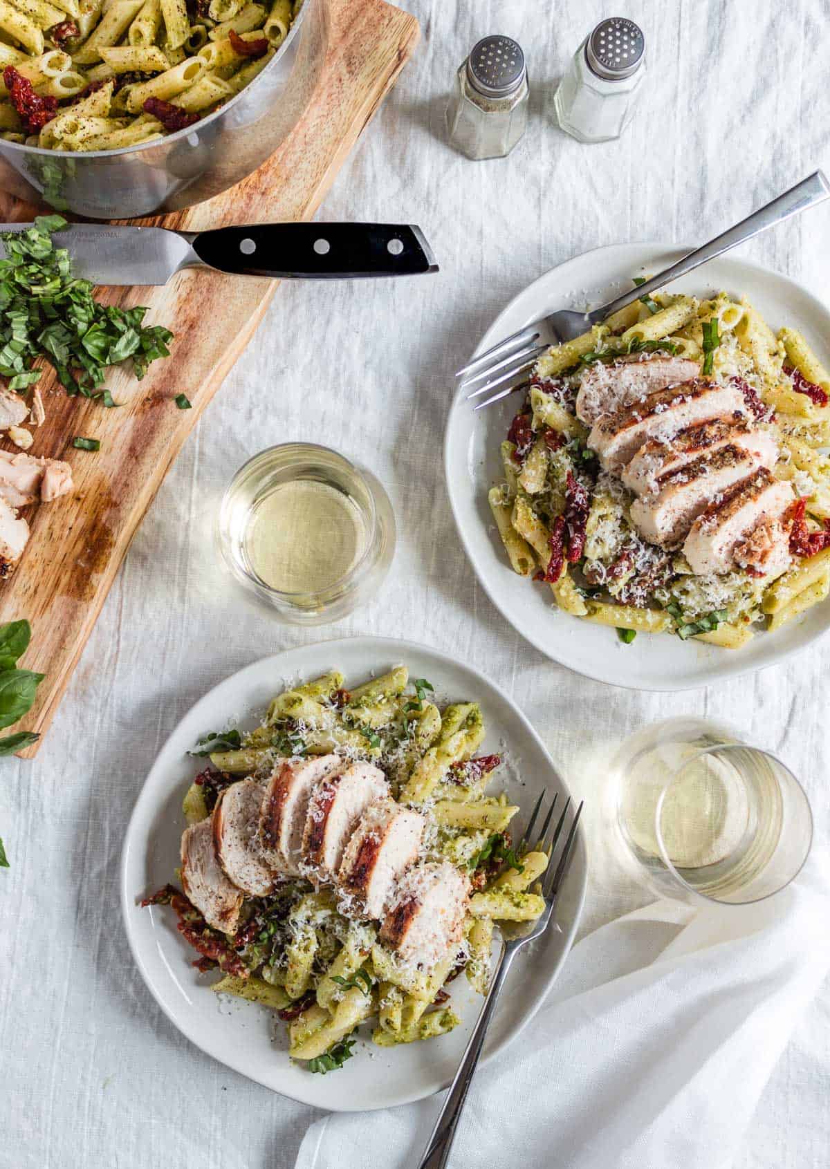 Two plates of creamy pesto pasta topped with grilled chicken, sun-dried tomatoes, and grated parmesan. Two glasses of white win sit nearby as well as a pot of pasta seconds and a serving board with sliced grilled chicken.