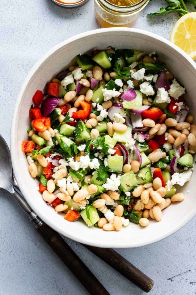 A large bowl of Mediterranean Bean Salad with serving utensils, lemon, and a jar of leftover vinaigrette off to the sides.