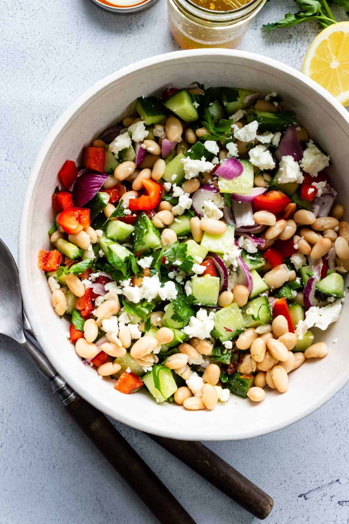 A large bowl of Mediterranean Bean Salad with serving utensils, lemon, and a jar of leftover vinaigrette off to the sides.