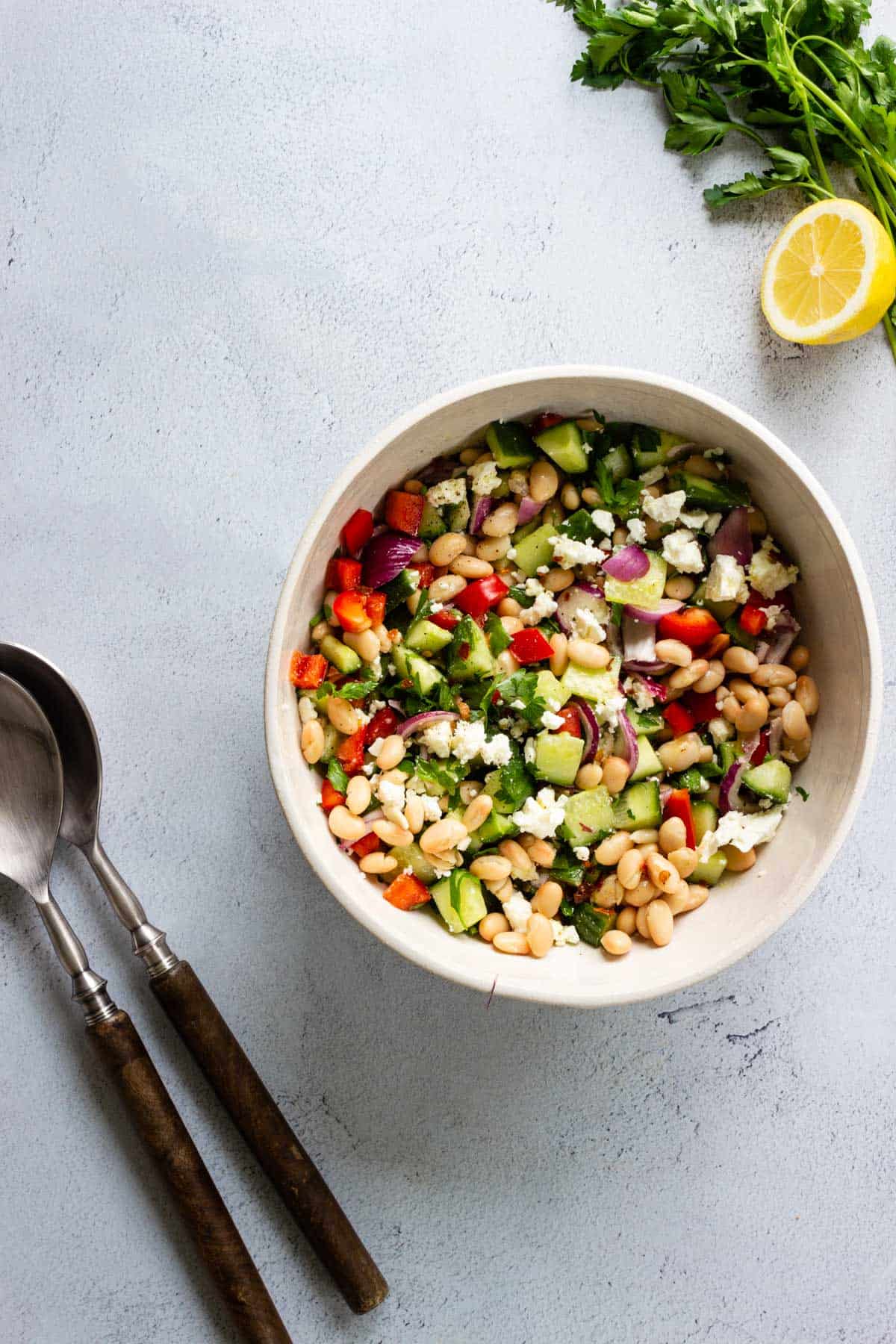 A bowl of Mediterranean Bean Salad and serving utensils are sitting on a gray countertop.