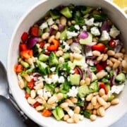 A large bowl of Mediterranean Bean Salad with serving utensils, lemon, and a jar of leftover vinaigrette off to the sides.